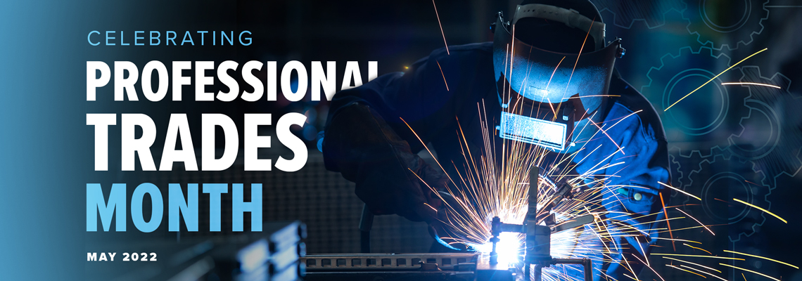 Celebrating Professional Trades Month, May 2022. Metal fabrication worker in a blue jumpsuit wearing a welding helmet and using a welding torch to join a clamped metal piece while sparks spread.