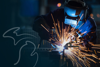 A welder clad in protective gear including a welding mask, gloves, and a jumpsuit, operating a torch amidst a shower of sparks.