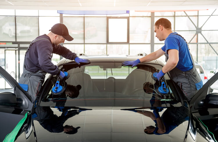 Two white automotive workers wearing overalls and using suction cups to install a new windshield on a car in a brightly-lit shop.