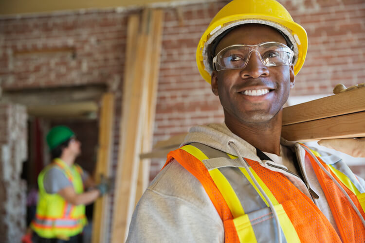 A smiling Black construction tradesman wearing a hard hat, safety glasses, and an orange safety vest carrying lumber on a construction site.